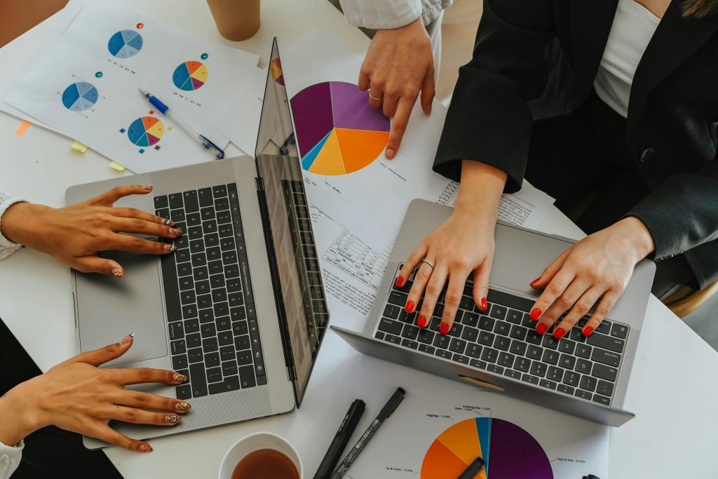 pexels-photo-8145342-8145342 Top view of women working together on laptops with business charts at a white office table.